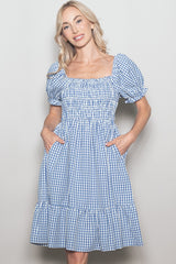 woman wearing a blue and white gingham puff sleeve dress with a smocked bodice and ruffled hem, posing with hands in pockets