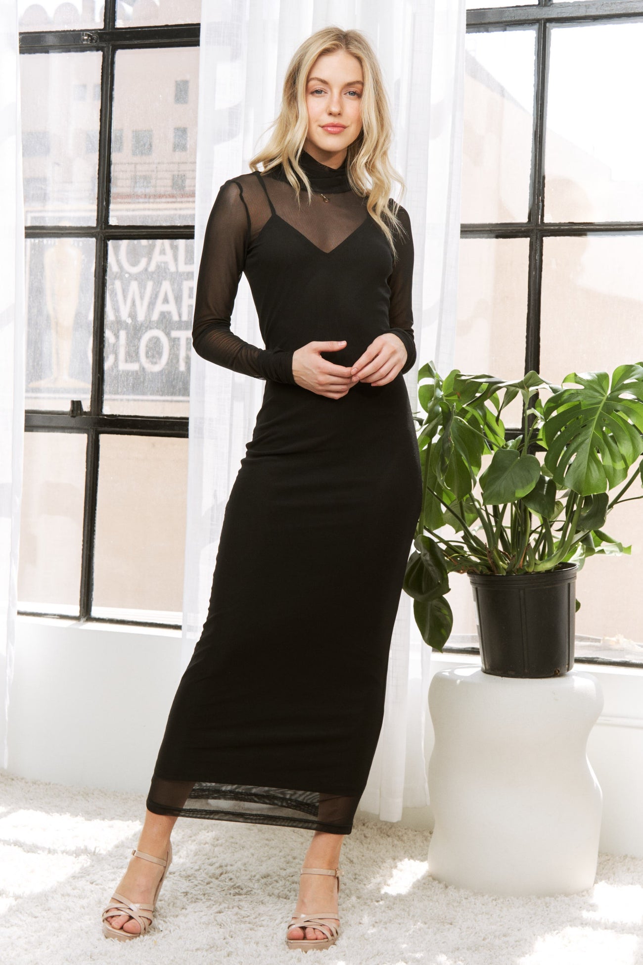 woman wearing a black long sleeve fitted dress with mesh detailing, standing next to a potted plant