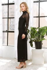 woman wearing a black sheer long sleeve maxi dress with a high neckline, standing next to a potted plant