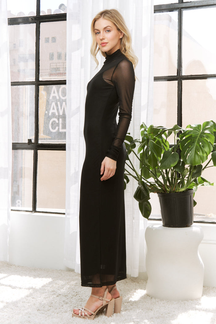 woman wearing a black sheer long sleeve maxi dress with a high neckline, standing next to a potted plant