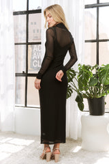 woman wearing a black long sleeve sheer maxi dress with a fitted silhouette, standing near a window