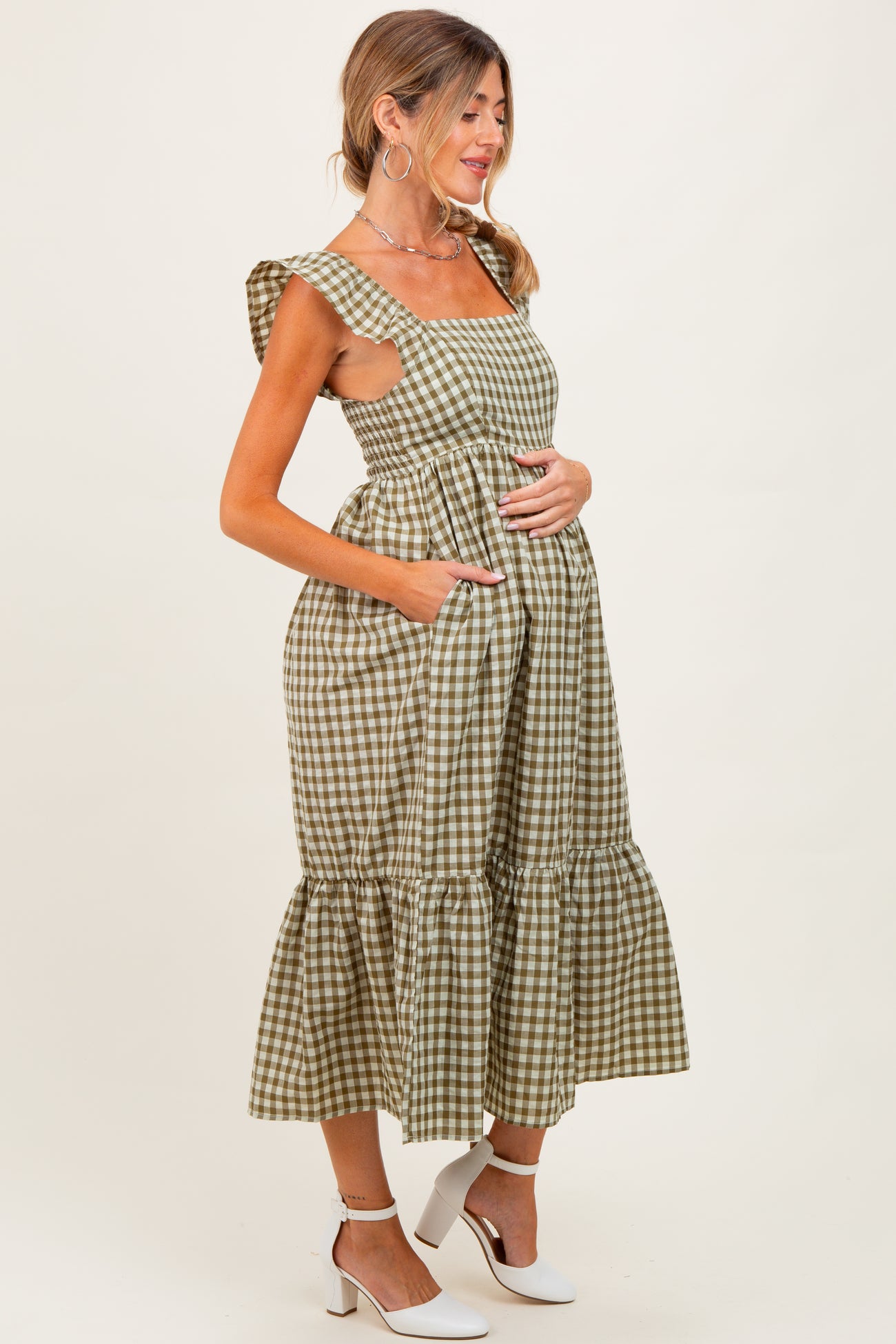 woman wearing a green and beige checkered maternity dress with ruffle straps and a tiered skirt, holding her baby bump