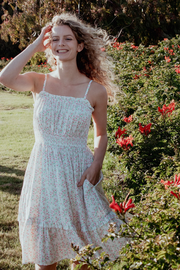 woman wearing a white floral sundress with thin straps and a tiered hem, posing in a garden with red flowers