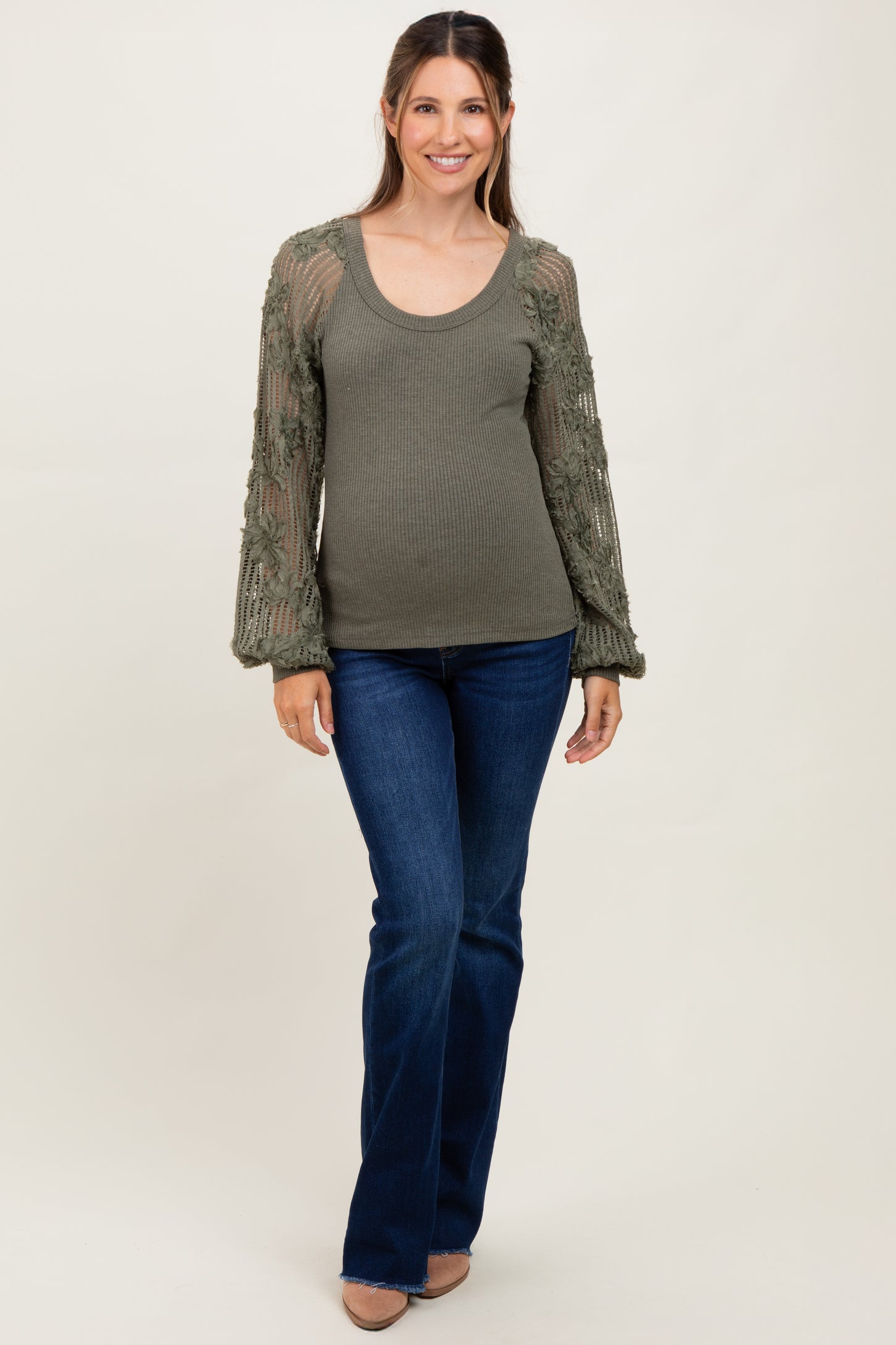 woman wearing a green ribbed maternity top with floral crochet sleeves and blue flared jeans, showing her baby bump