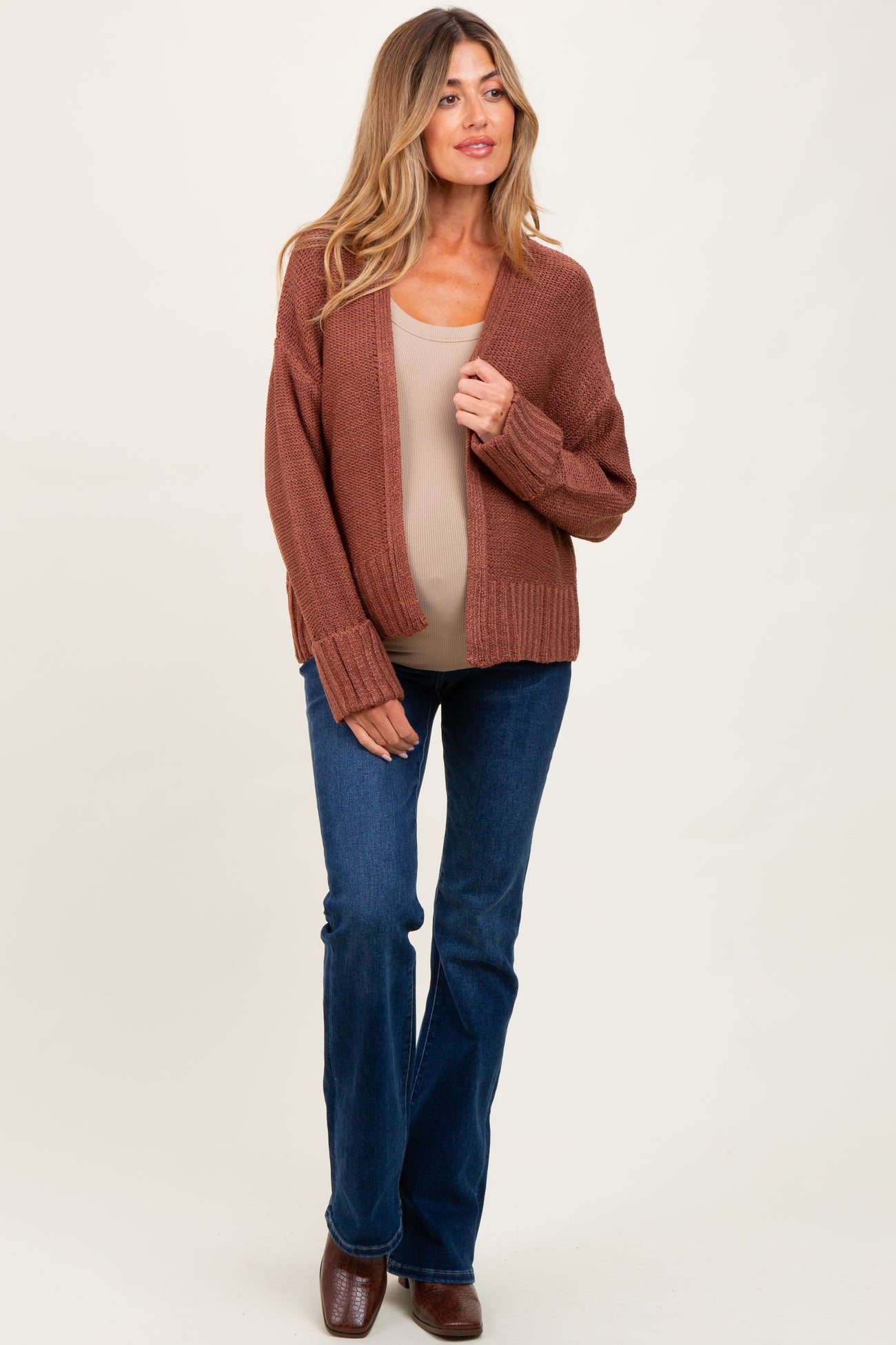 woman wearing a rust-colored maternity cardigan, fitted beige top, and flared jeans, showcasing her baby bump