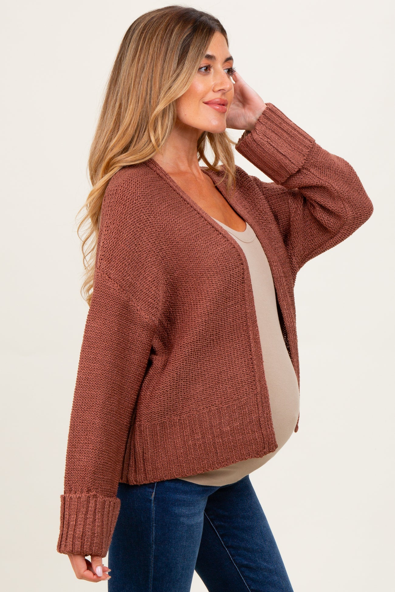 woman wearing a rust-colored open-knit maternity cardigan over a taupe top and showing her baby bump, styled with jeans