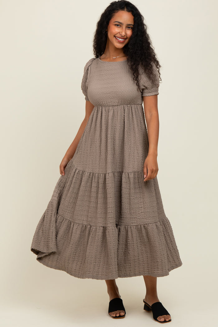 woman wearing a taupe tiered puff sleeve maxi dress with a textured fabric and black slide sandals
