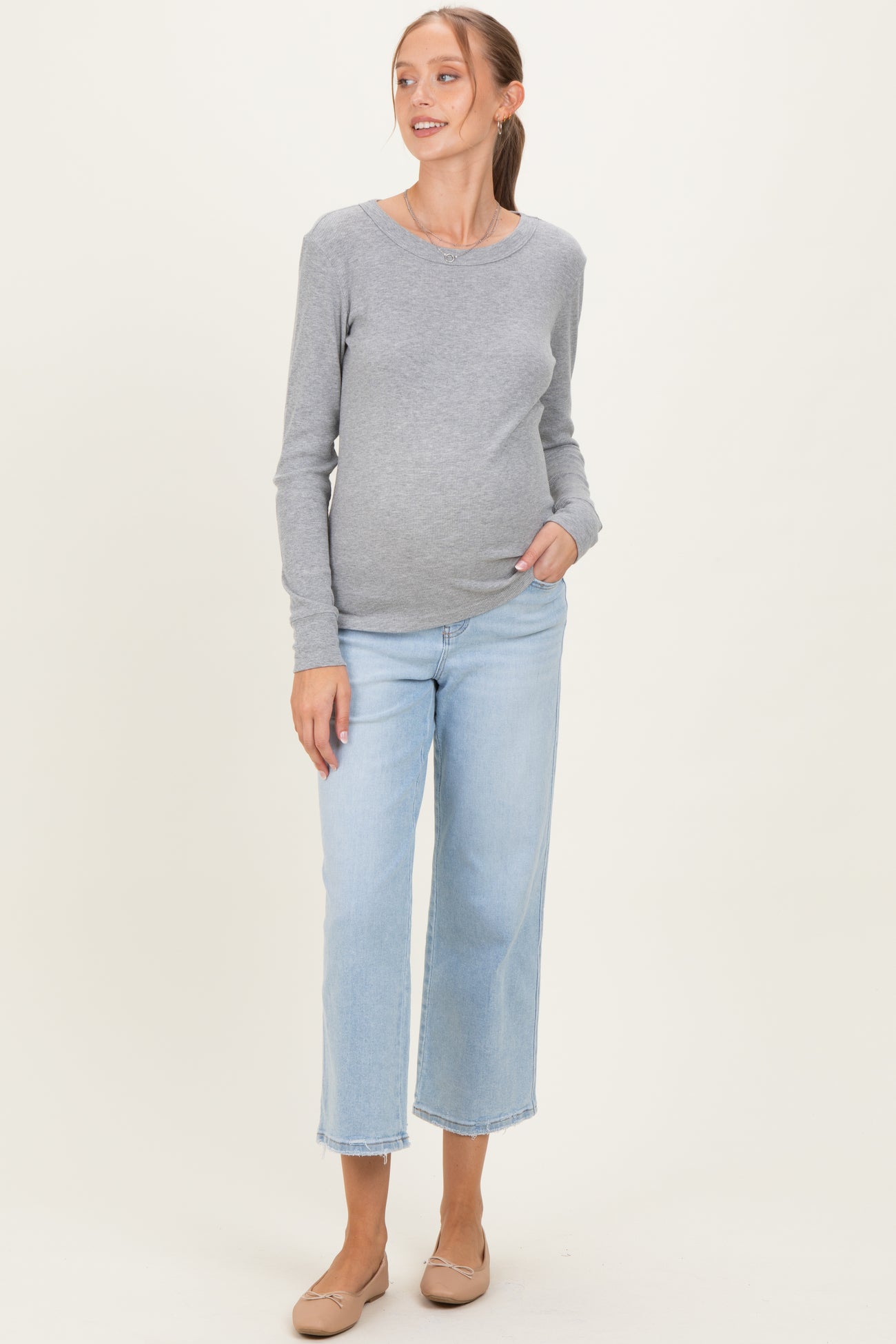 woman wearing a gray long sleeve maternity top and light blue cropped jeans, gently cradling her baby bump