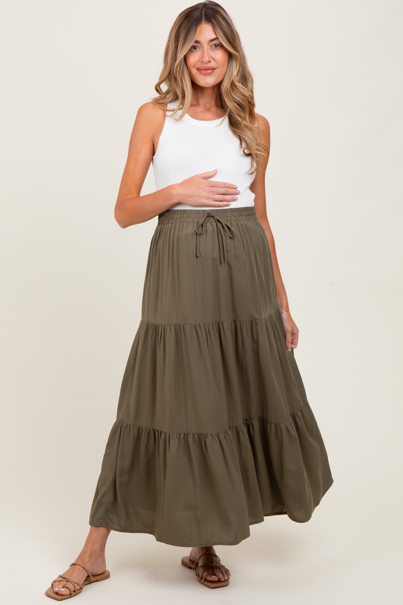 woman wearing a white tank top and an olive green tiered maternity skirt, holding her baby bump