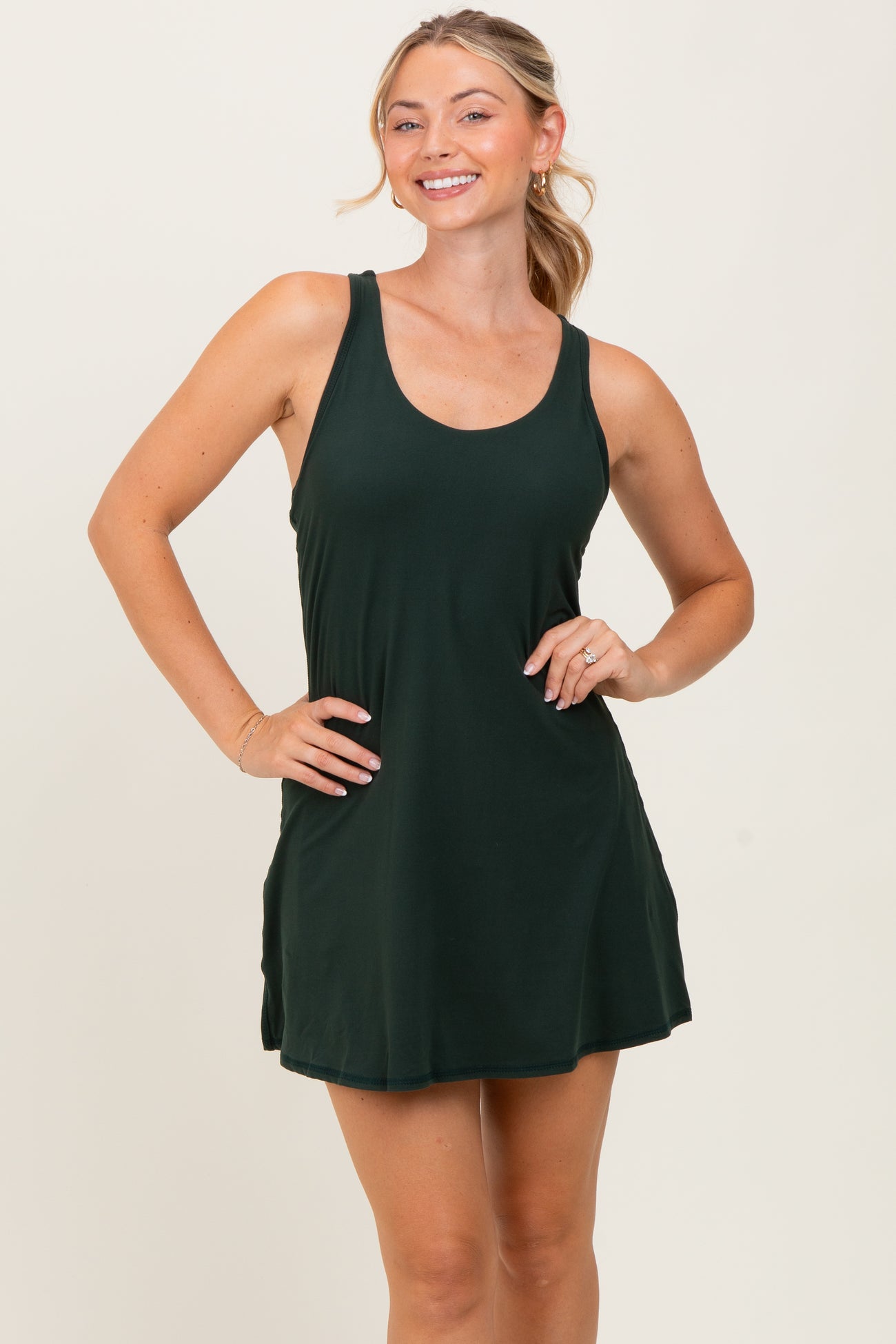 woman wearing a dark green tank dress with a flared hem, smiling and posing with one hand on her hip