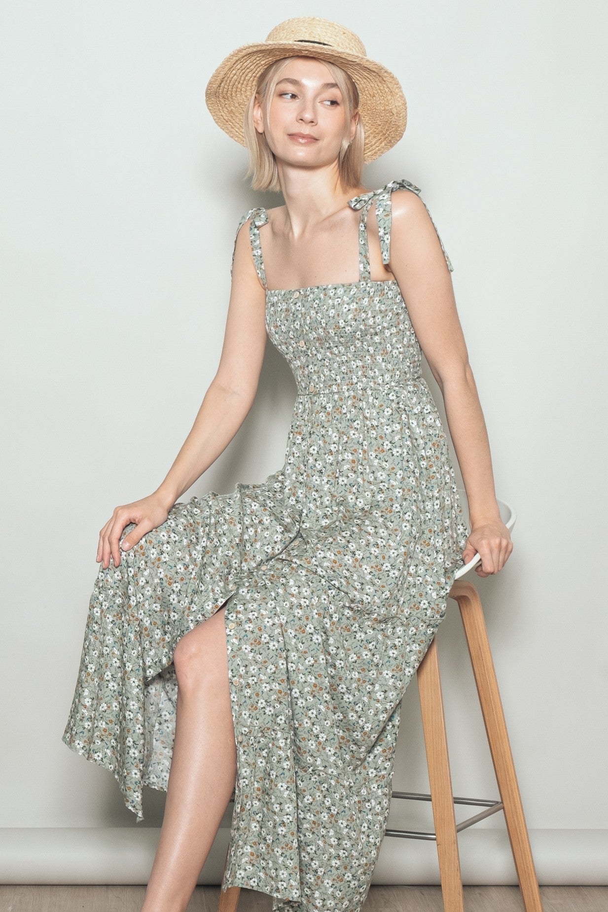 woman wearing a green floral maxi dress with tie straps and a straw hat, sitting on a stool