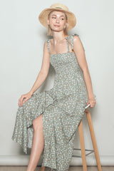 woman wearing a green floral maxi dress with tie straps and a straw hat, sitting on a stool