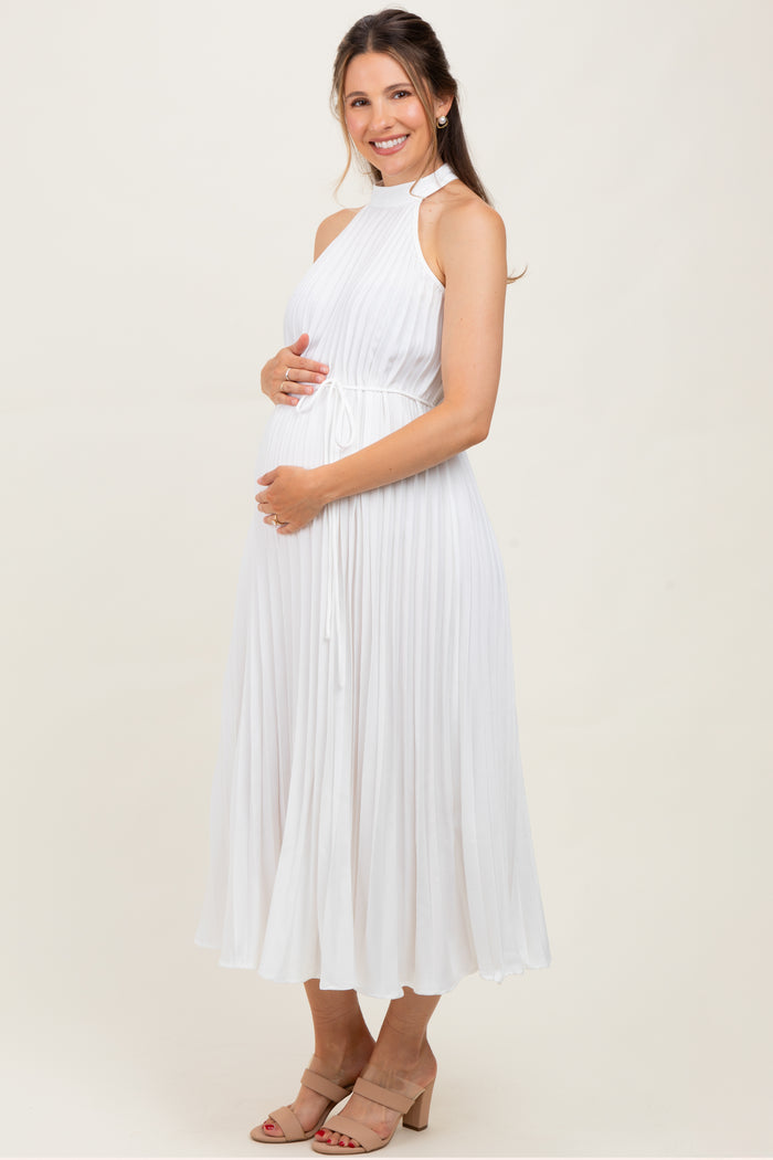 woman wearing a white pleated maternity dress with a high neckline and holding her baby bump