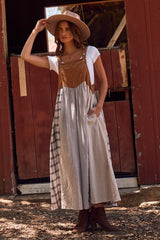 woman wearing a brown corduroy and checked overall dress with side pockets, white shirt underneath, and hat, standing near a barn