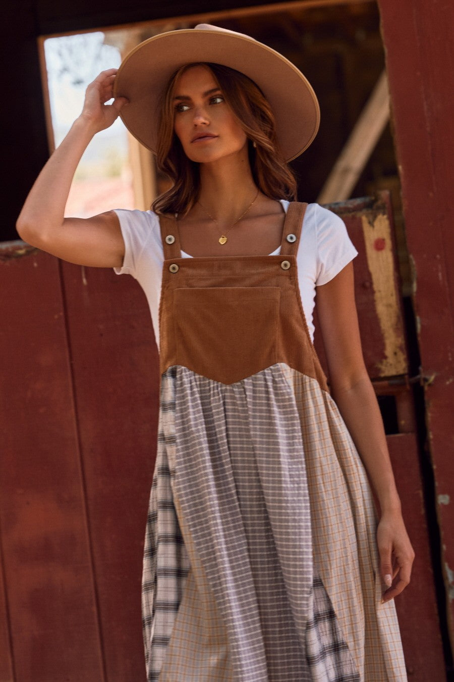 woman wearing a brown and plaid jumper dress over a white t-shirt and a wide-brimmed hat