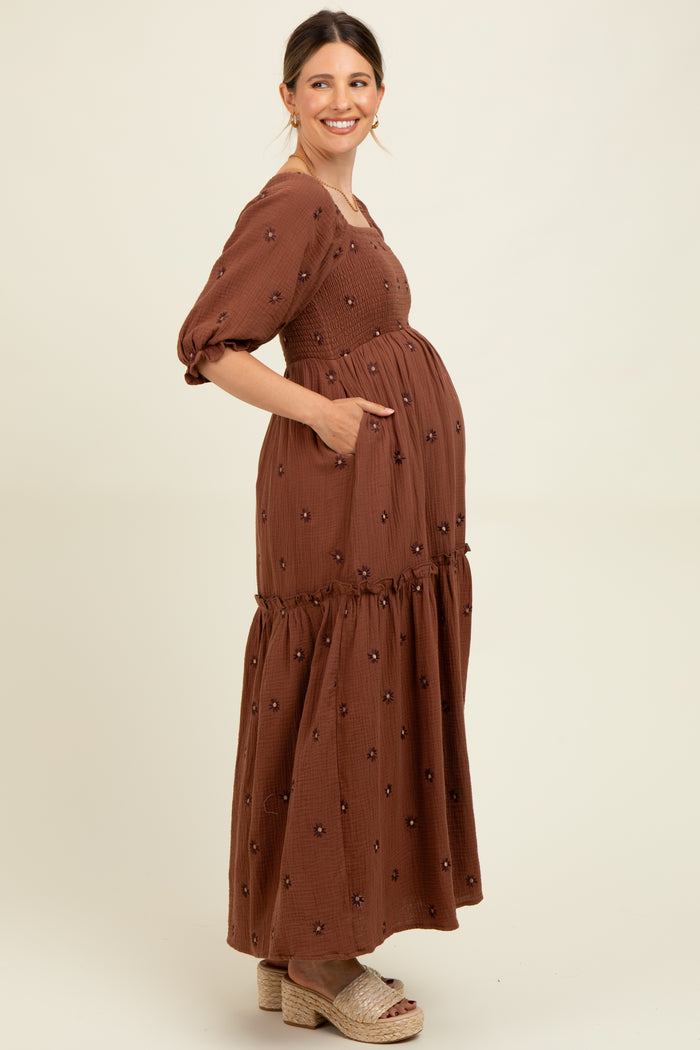woman wearing a brown maternity maxi dress with puff sleeves and floral embroidery, showing her baby bump and pockets