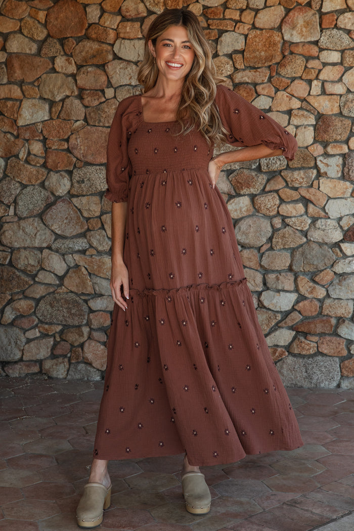 woman wearing a brown maternity maxi dress with puff sleeves and floral embroidery, showing her baby bump