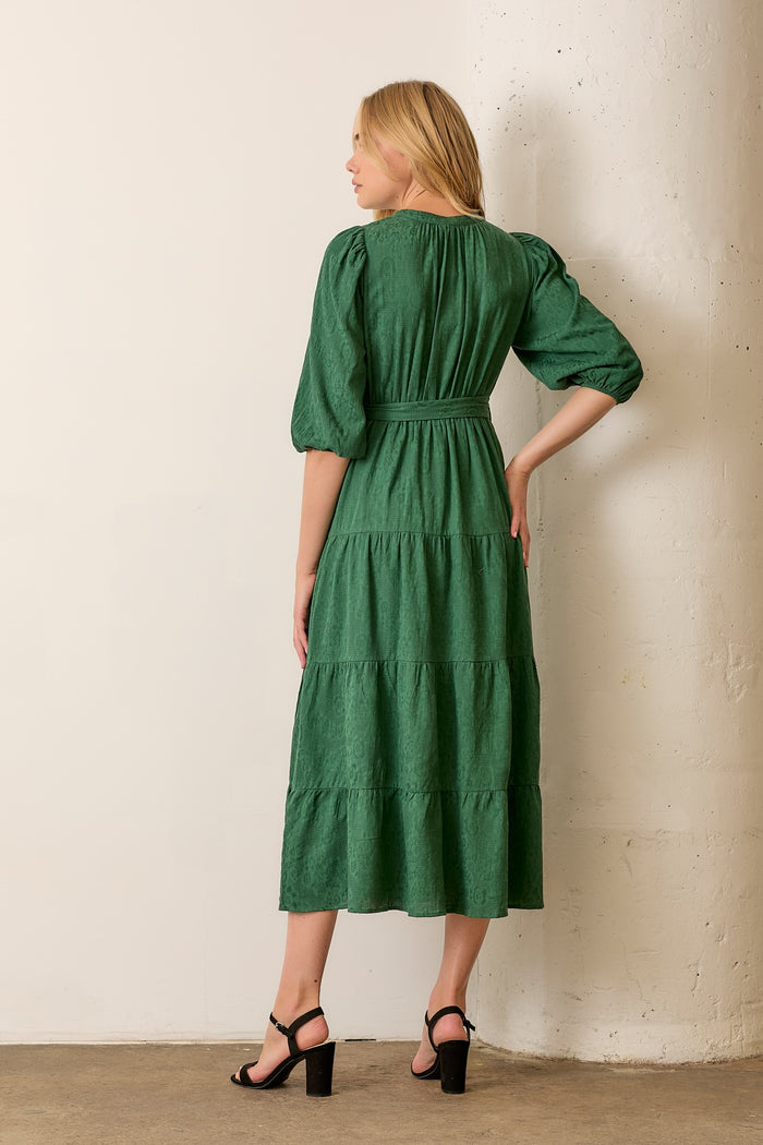 woman wearing a green tiered midi dress with puff sleeves and a waist tie, standing against a white wall