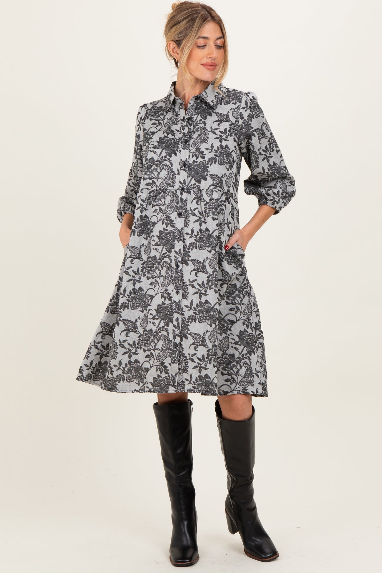 woman wearing a black and white floral print maternity dress with buttons and pockets, standing in tall black boots