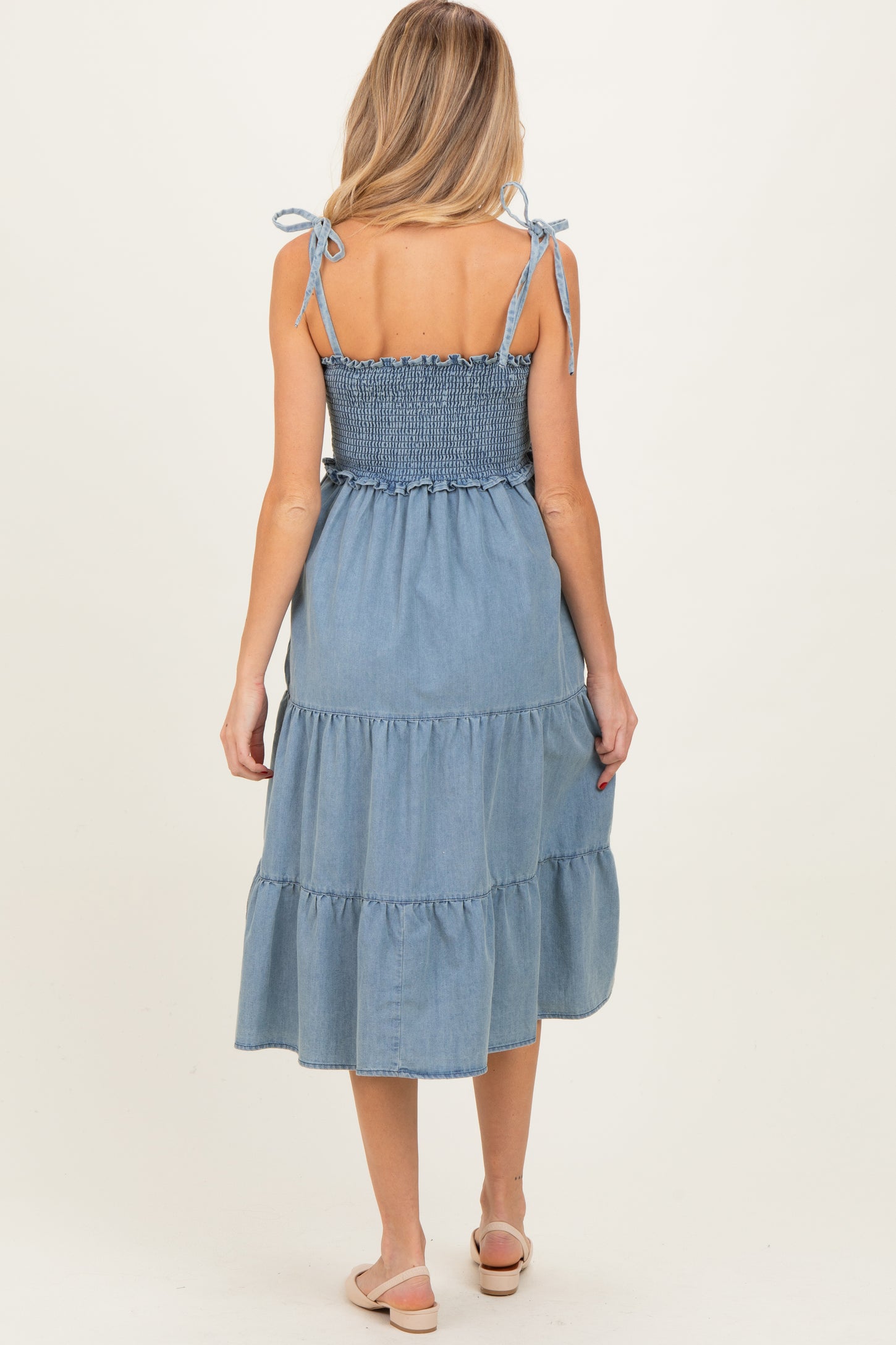 woman wearing a light blue denim smocked maternity dress with tiered ruffles and tie straps, showing her back