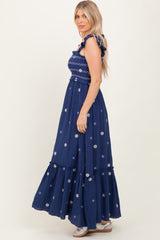 woman wearing a navy maxi dress with floral embroidery and ruffled straps, standing in profile against a light background