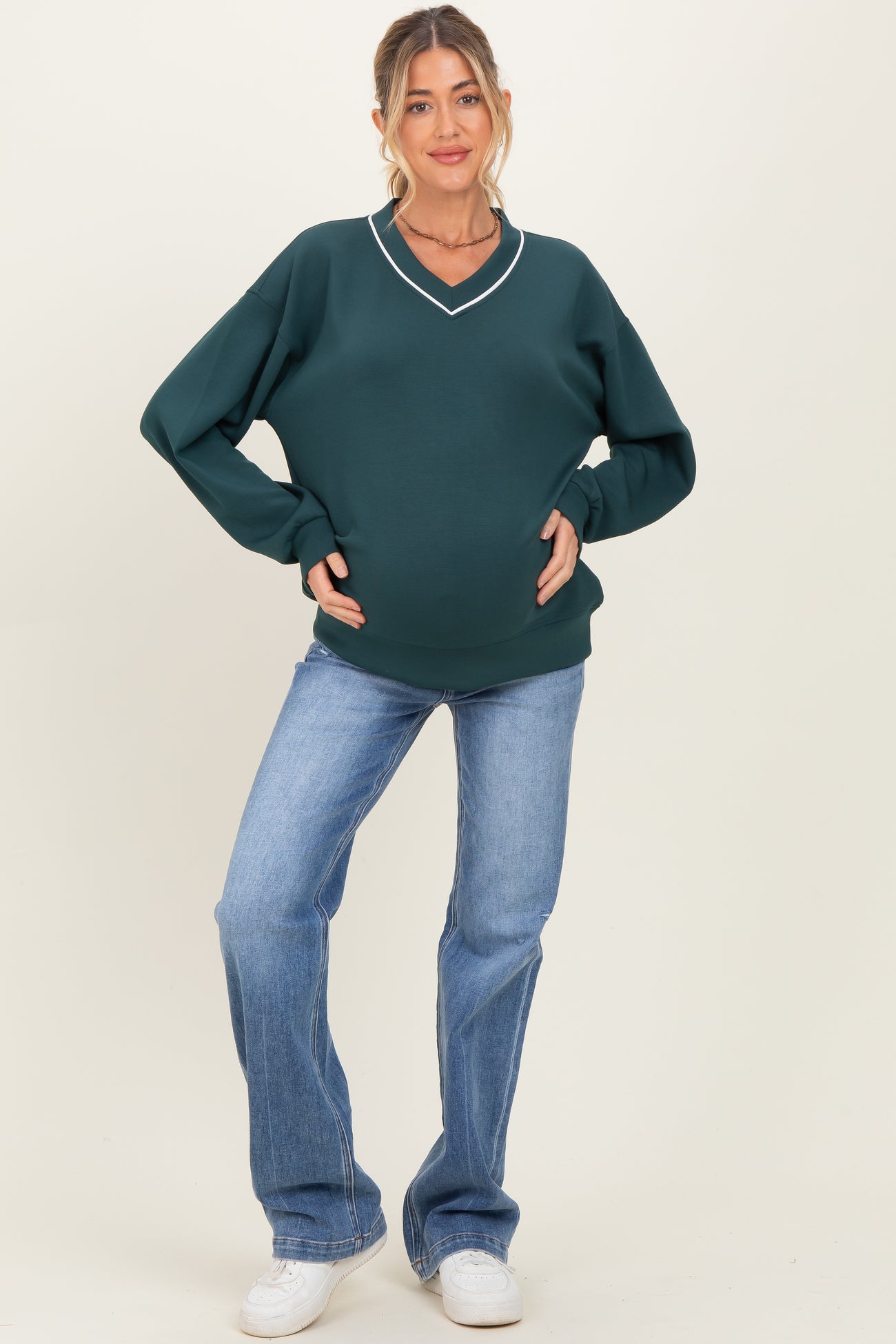 woman wearing a dark green maternity sweatshirt with a v-neckline and holding her baby bump while standing in light blue jeans