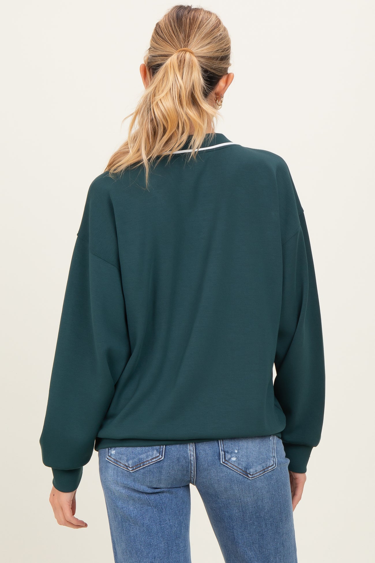 woman wearing a dark green long sleeve maternity sweatshirt with white detailing and blue jeans, showing a relaxed fit from the back