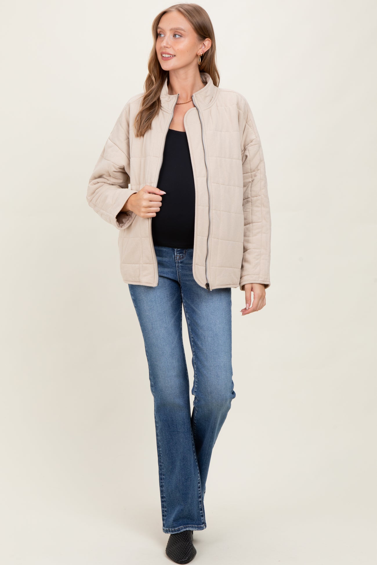 woman wearing a beige quilted maternity jacket over a black top and blue jeans, with a visible baby bump
