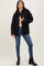 woman wearing a black quilted jacket, light pink top, and blue jeans standing in front of a neutral background