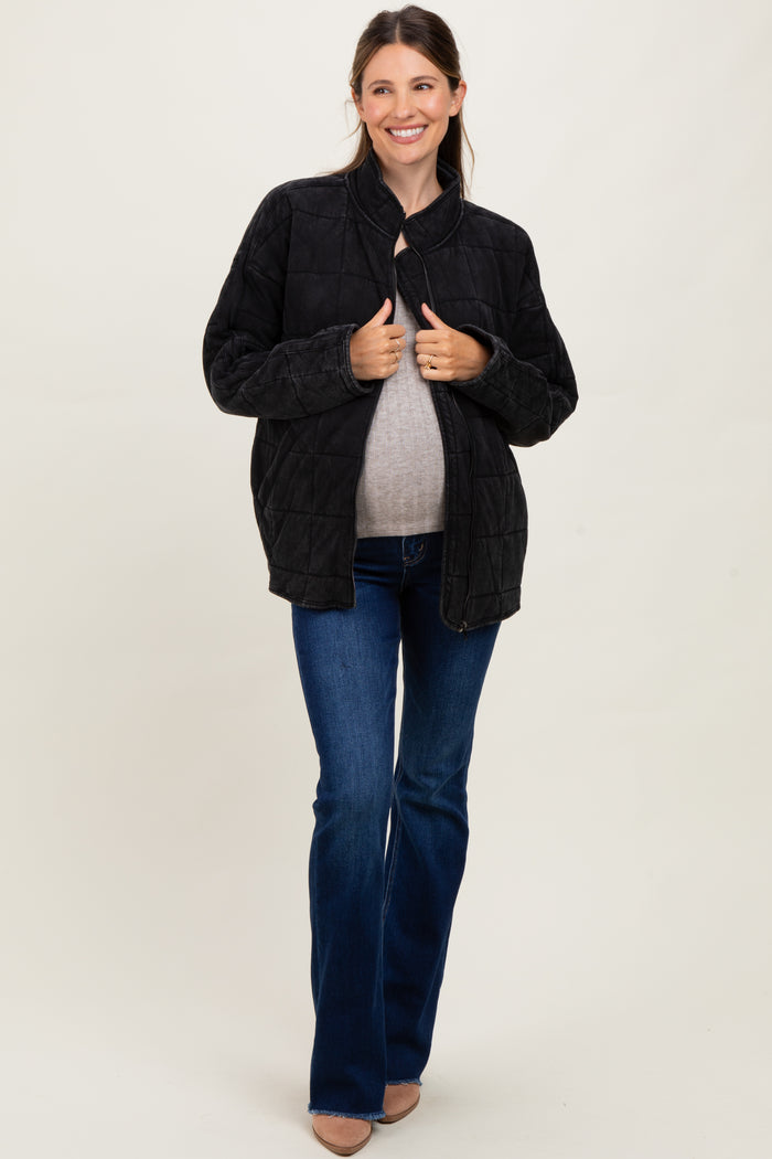 woman wearing a black quilted maternity jacket and a gray top, holding her baby bump while standing in flared jeans