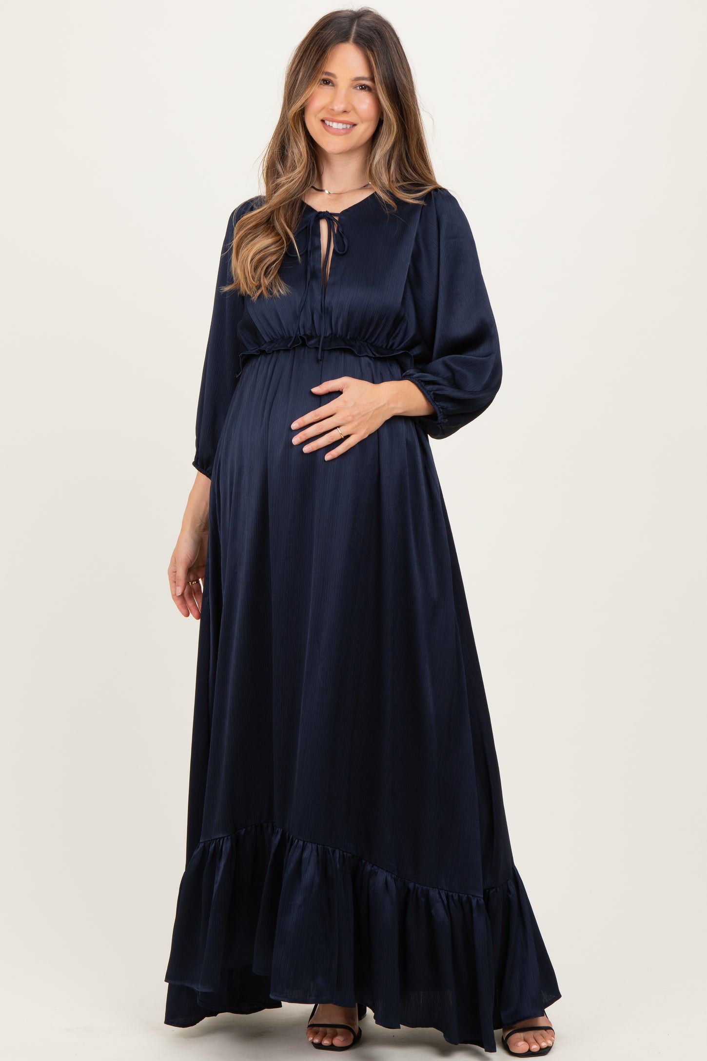 woman wearing a navy blue maternity maxi dress with a tie neck and ruffled hem, cradling her baby bump