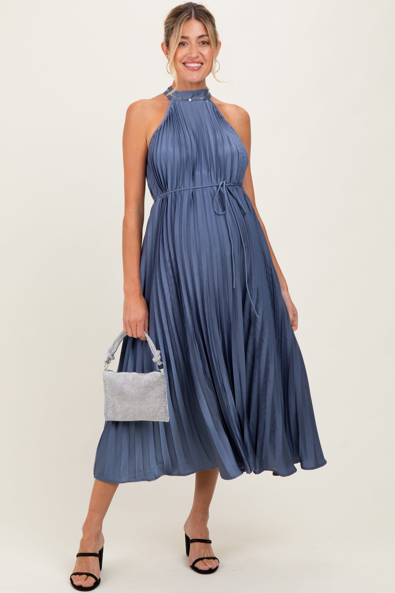 woman wearing a navy blue pleated maternity dress with a halter neckline and a cinched waist, holding a handbag