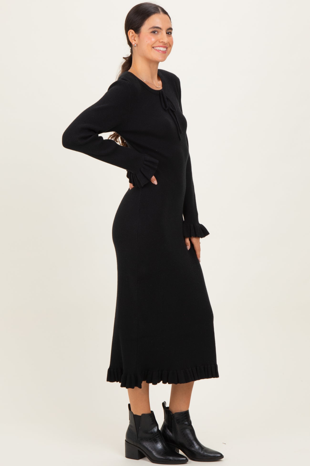 woman wearing a black ribbed long sleeve dress with ruffle details at the cuffs and hem, paired with black ankle boots