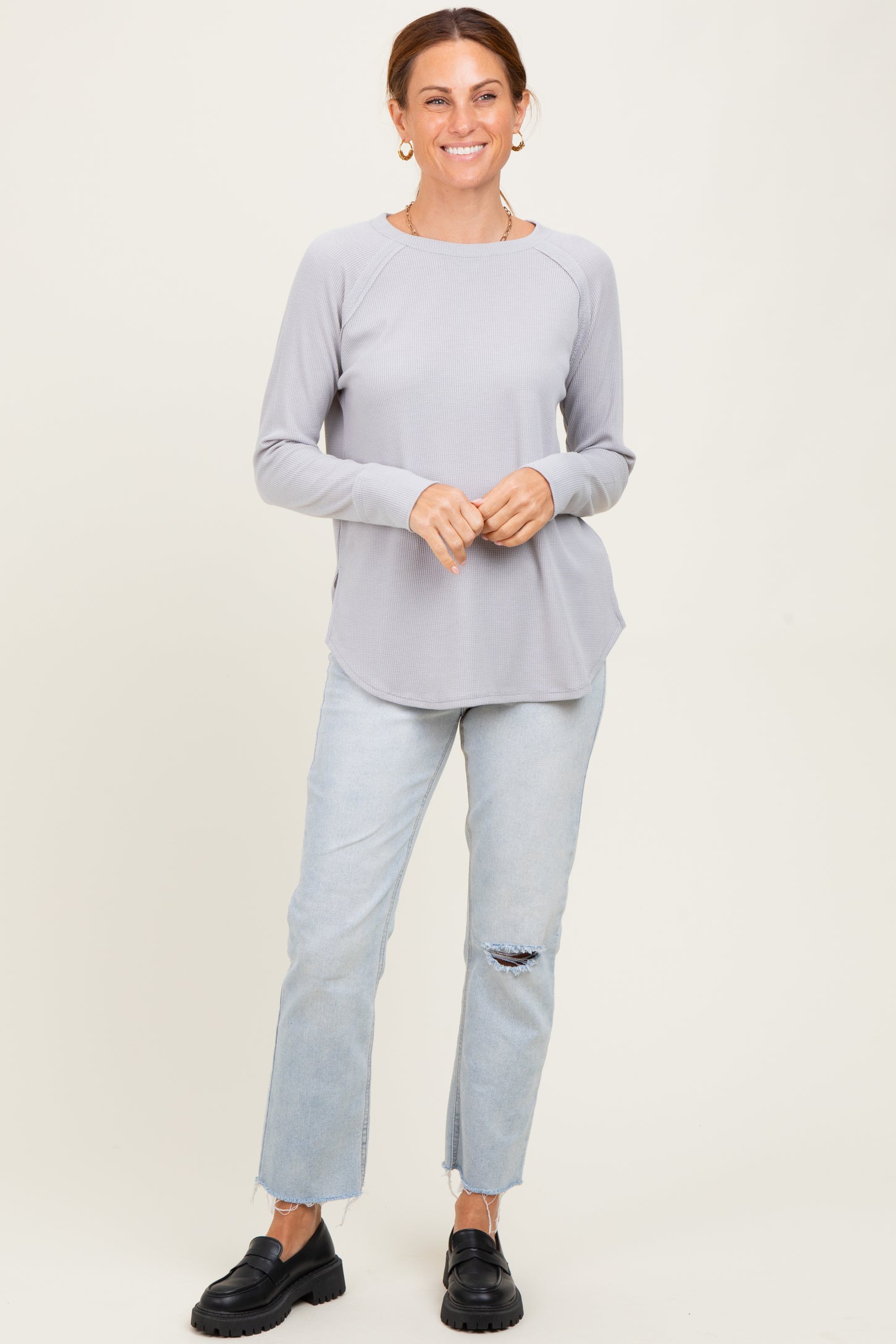 woman wearing a light gray long sleeve top with rounded hem and blue ripped jeans, standing and smiling