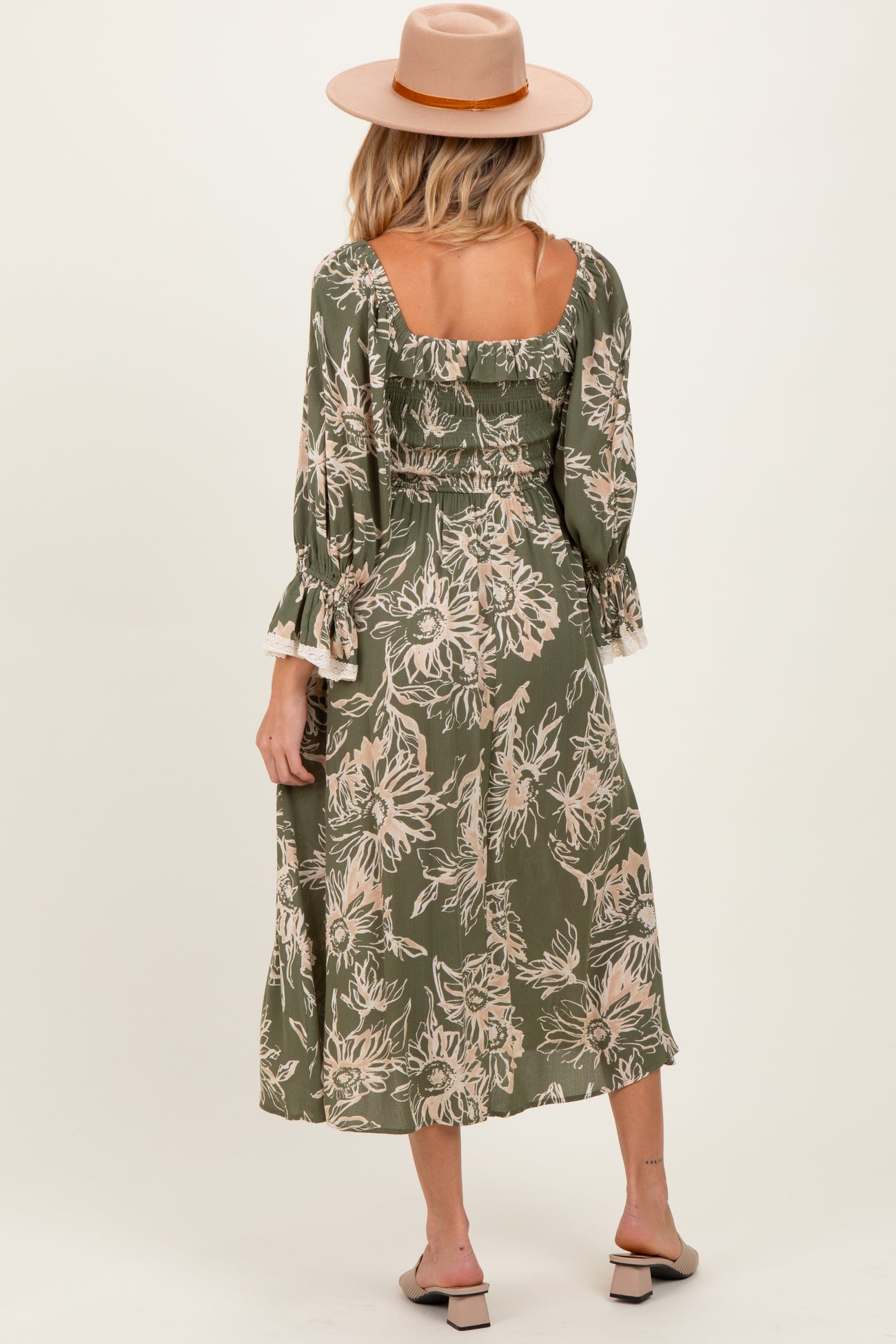woman wearing a green floral print maternity dress with ruffled sleeves and a square back, styled with a hat