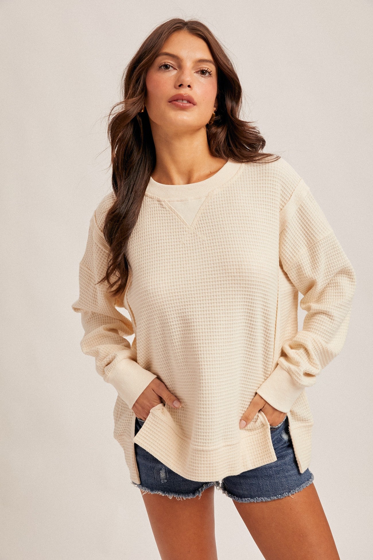 woman wearing a cream waffle knit long sleeve top with a relaxed fit and side slits paired with denim shorts