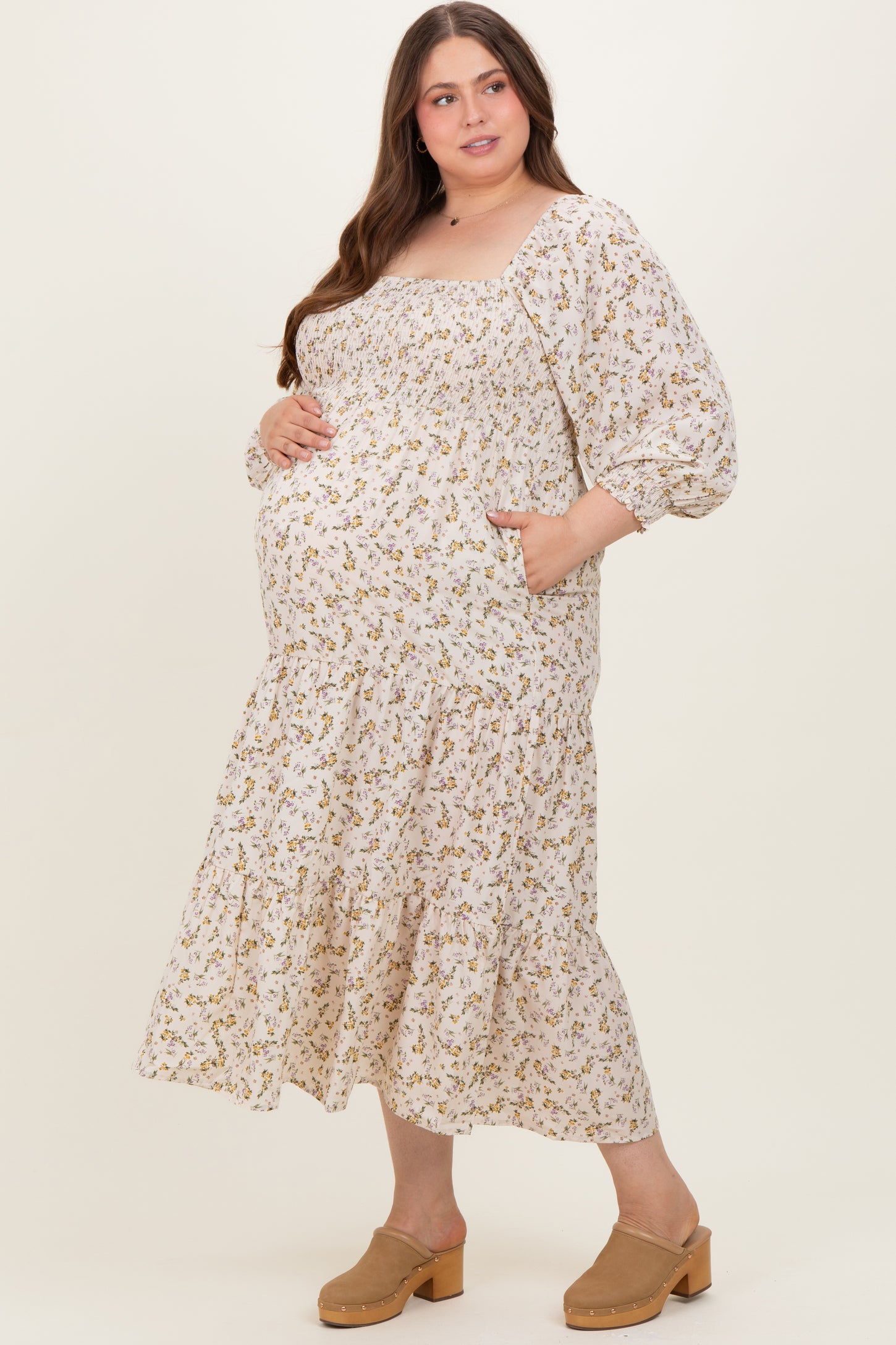 woman wearing a cream floral maternity dress with long puff sleeves and a tiered skirt, cradling her baby bump
