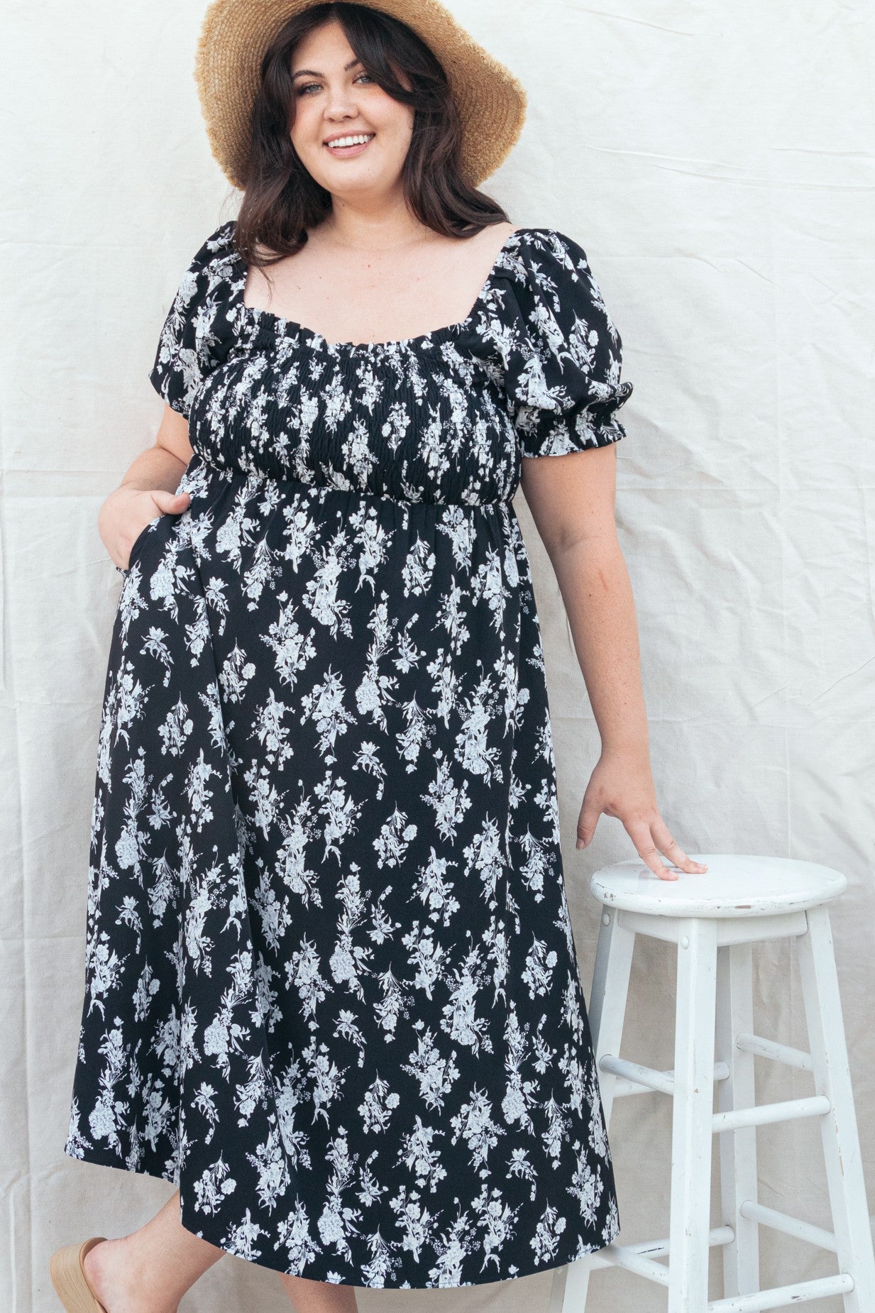 woman wearing a black floral print plus-size dress with puff sleeves and a cinched waist, smiling while standing beside a stool