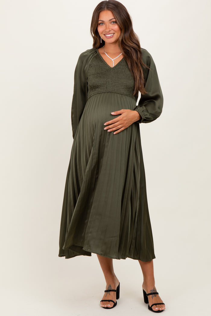 woman wearing an olive green pleated maternity dress with a smocked bodice and long sleeves, holding her baby bump