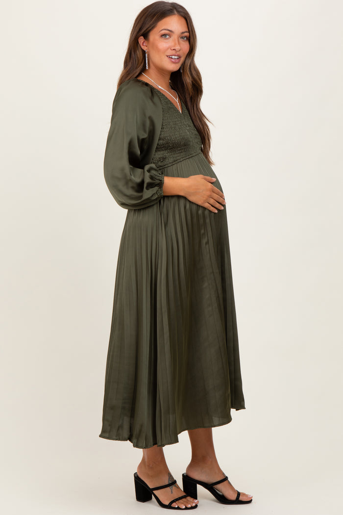 woman wearing an olive green maternity dress with smocked bodice and flowy skirt, gently cradling her baby bump