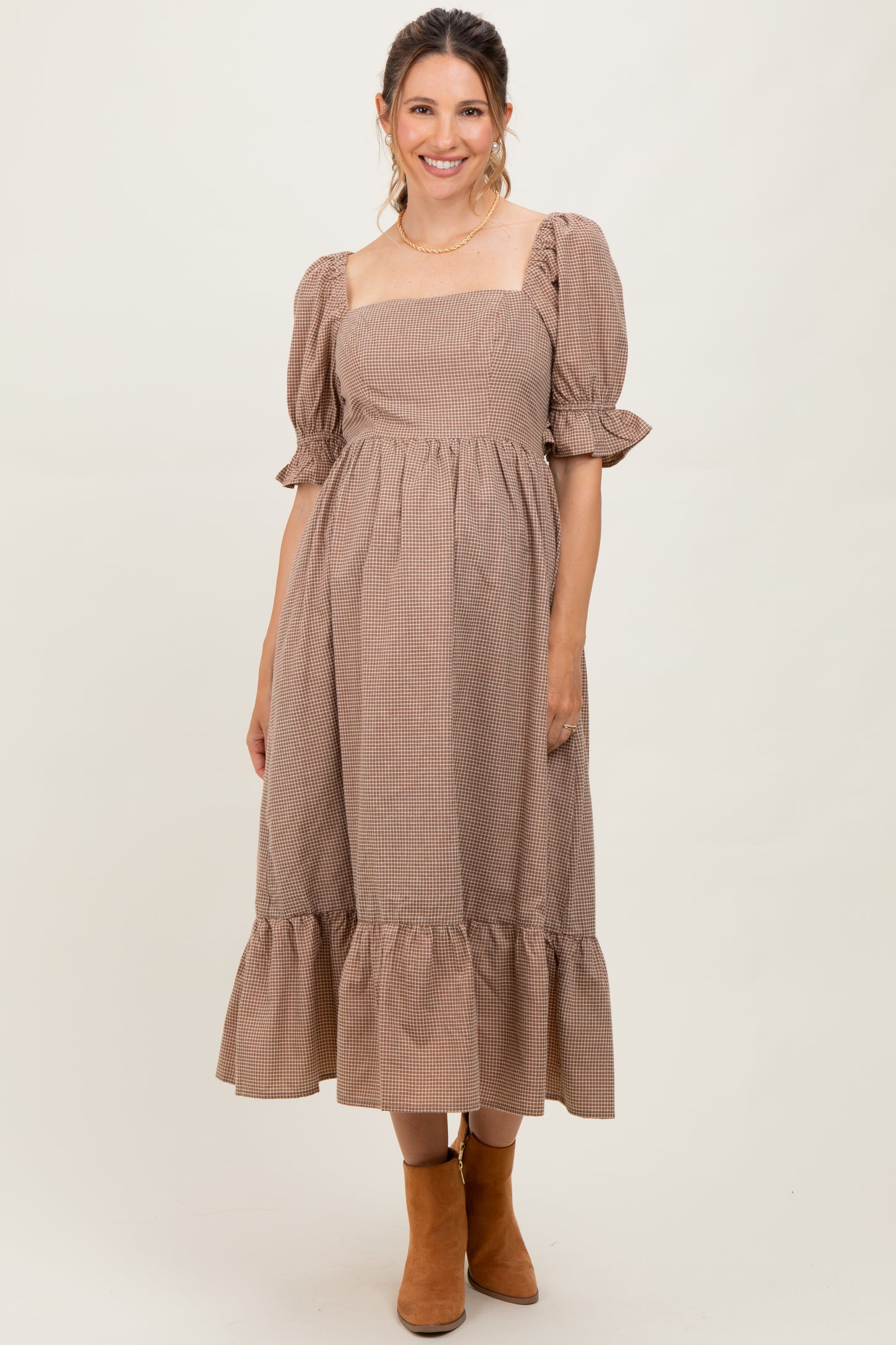 woman wearing a brown gingham maternity dress with short puff sleeves and a tiered skirt, posing with a slight smile