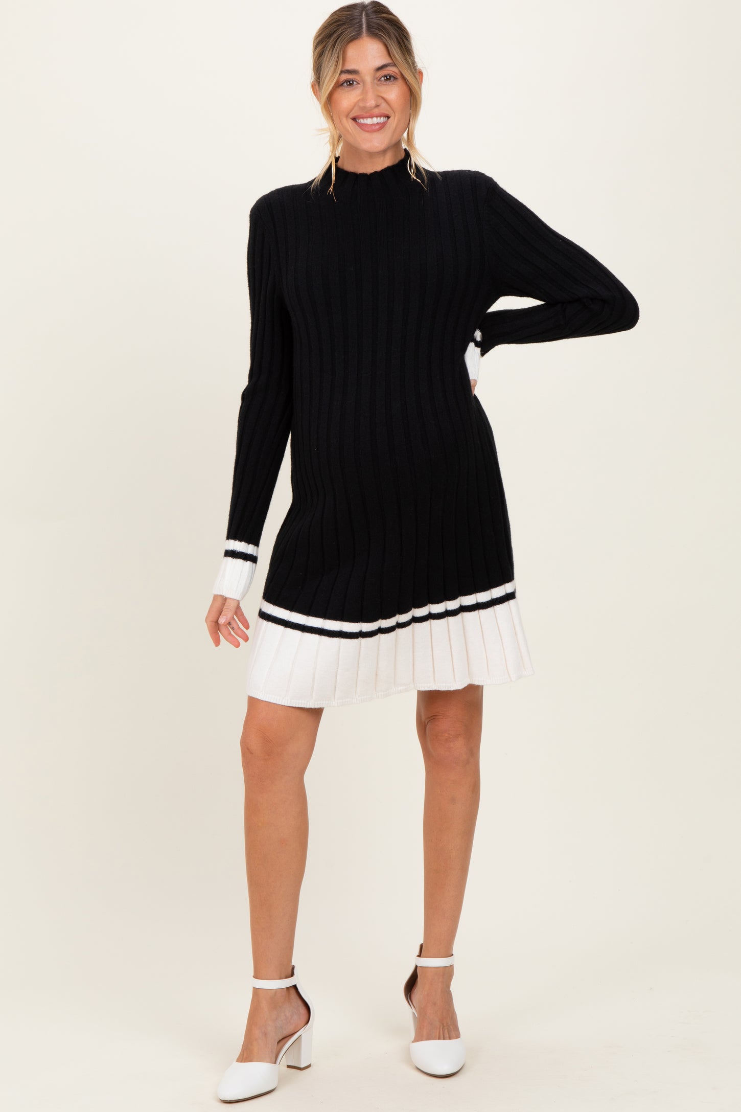 woman wearing a black ribbed maternity sweater dress with white pleated hem and holding her baby bump