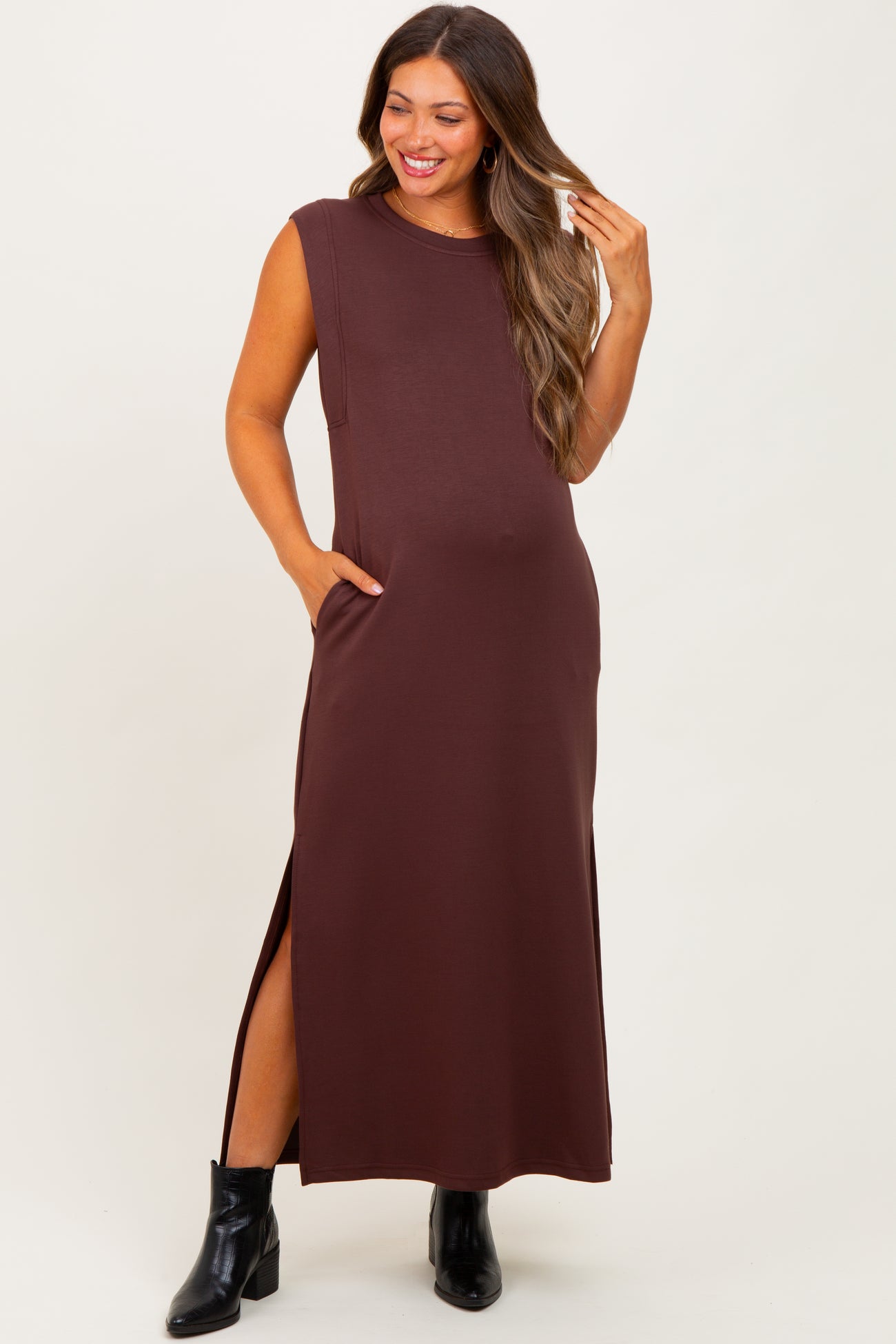 woman wearing a brown sleeveless maternity maxi dress with side slits and pockets, smiling and posing with one hand in her hair