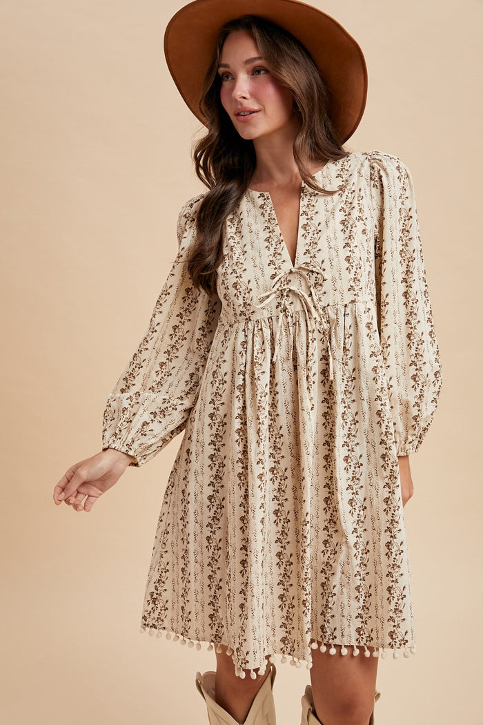 woman wearing a cream floral long sleeve dress with a tie detail and pom-pom trim, accessorized with a brown hat