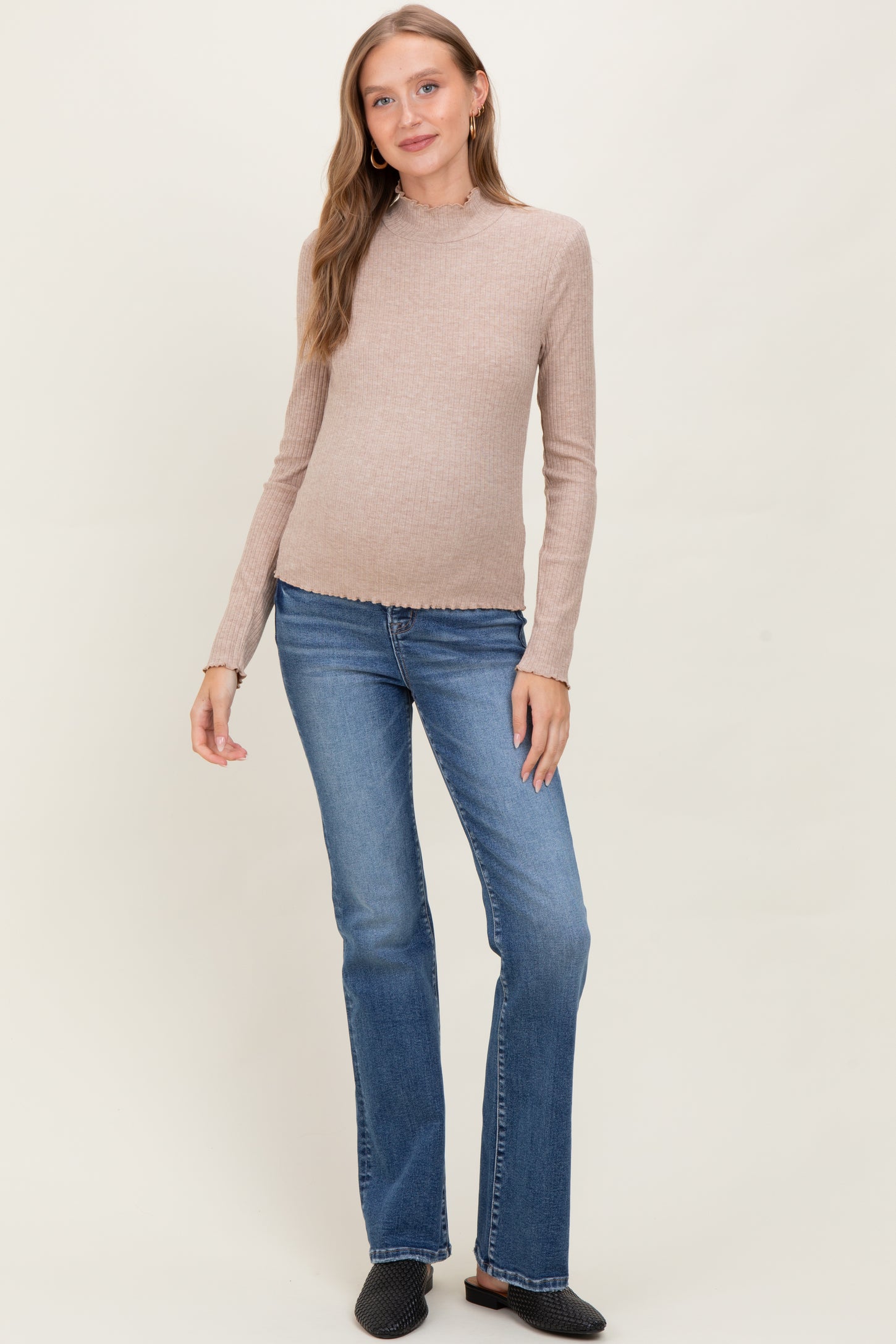 woman wearing a beige long sleeve maternity top with a scalloped hem and blue flared maternity jeans, showcasing a baby bump