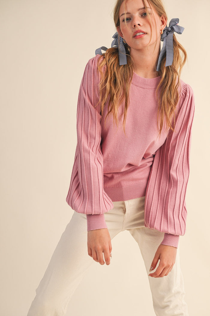 woman wearing a pink sweater with pleated sleeves and large bows in her hair, posing with one hand on her hip