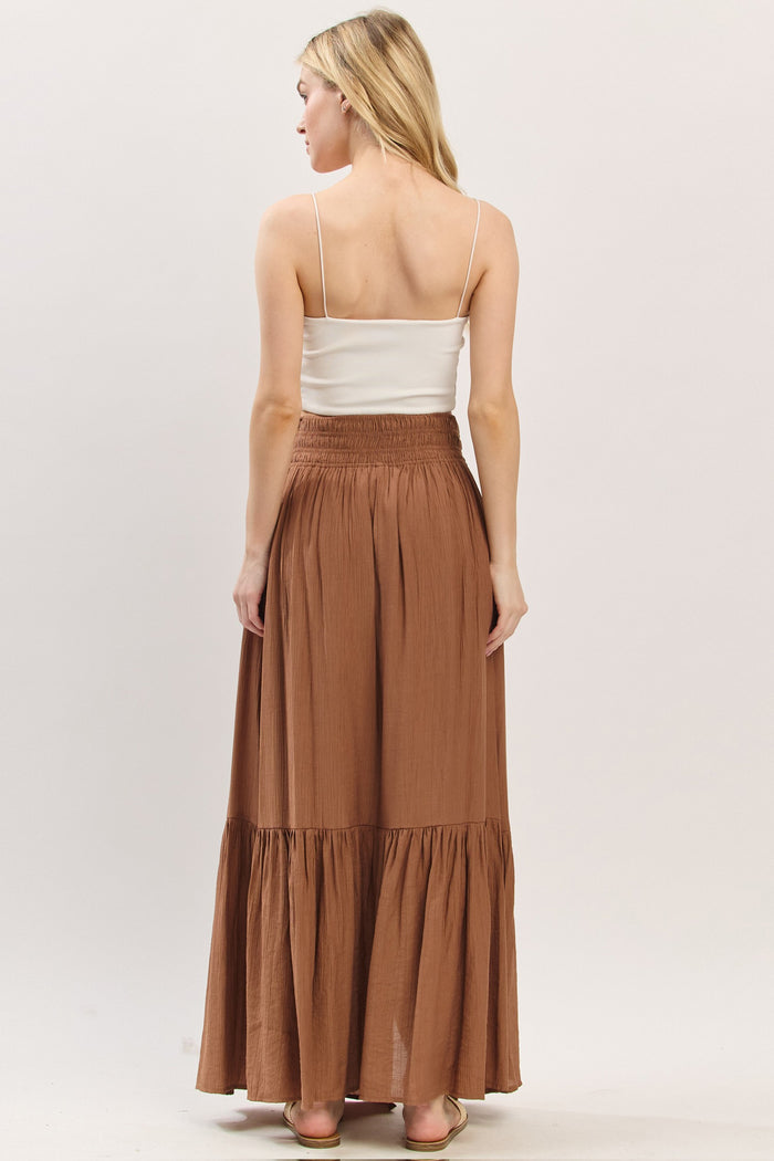 woman wearing a brown maxi skirt with a tiered hem and a white fitted camisole, viewed from the back