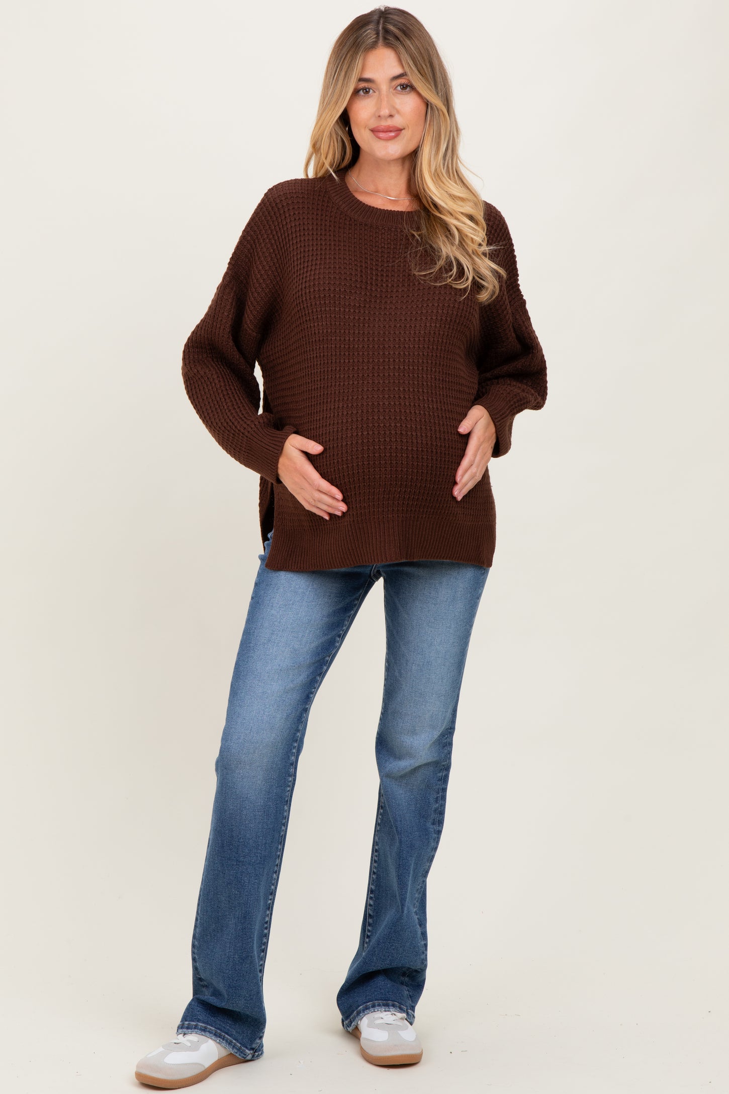 woman wearing a brown chunky knit maternity sweater and blue flared jeans, smiling while holding her baby bump