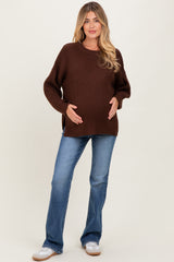 woman wearing a brown chunky knit maternity sweater and blue flared jeans, smiling while holding her baby bump