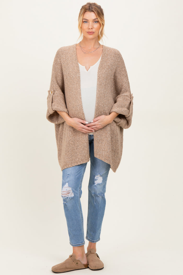 woman wearing a beige knitted maternity cardigan over a white top, holding her baby bump, paired with distressed jeans